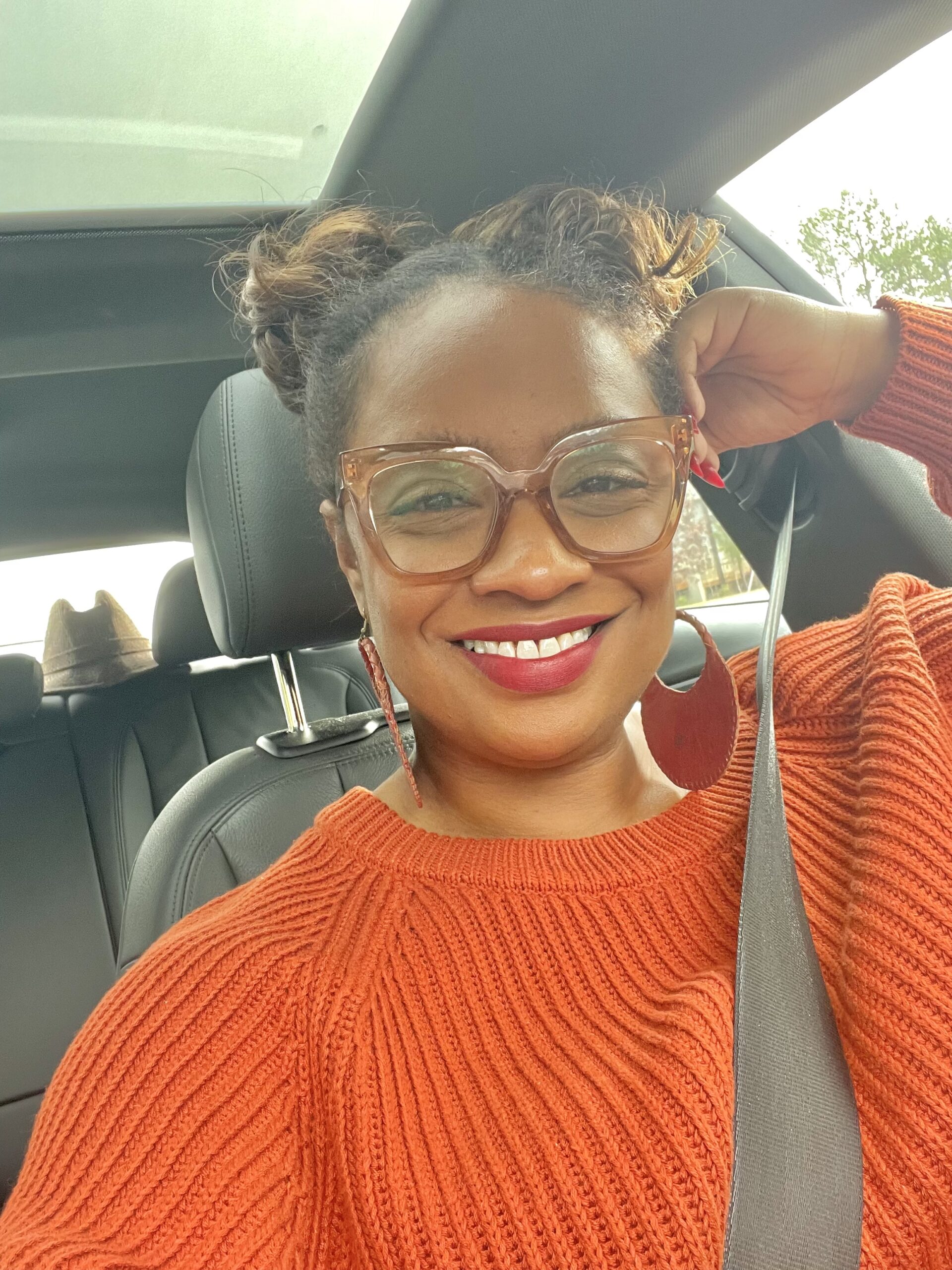 A smiling Black woman with brown skin, red lips and nails and a bright orange sweater